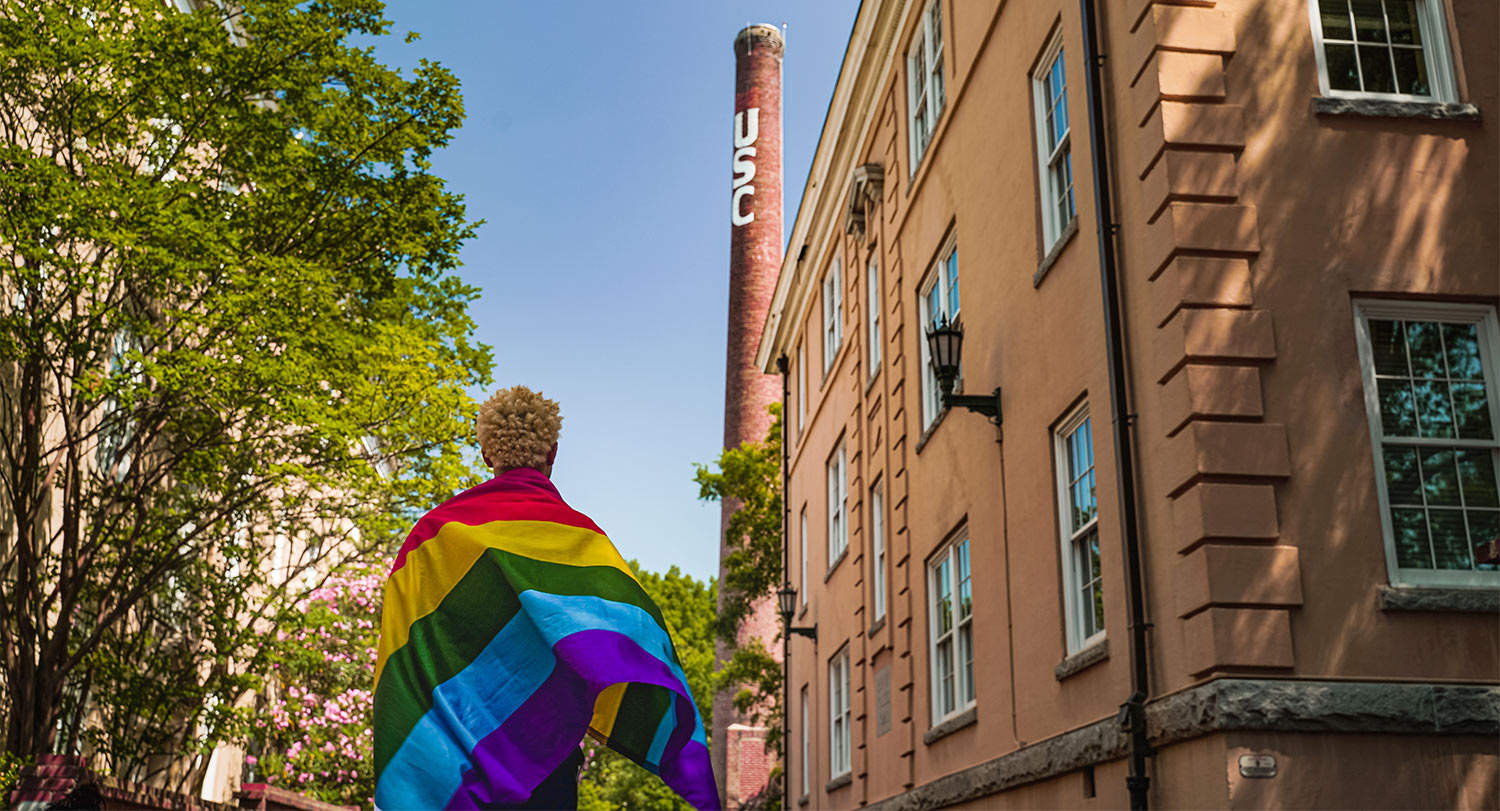 Student with a Pride flag wrapped around their shoulders with the USC smokestack in the background.