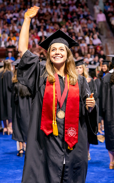 A student wearing robe and regalia waving to crowd at commencement.